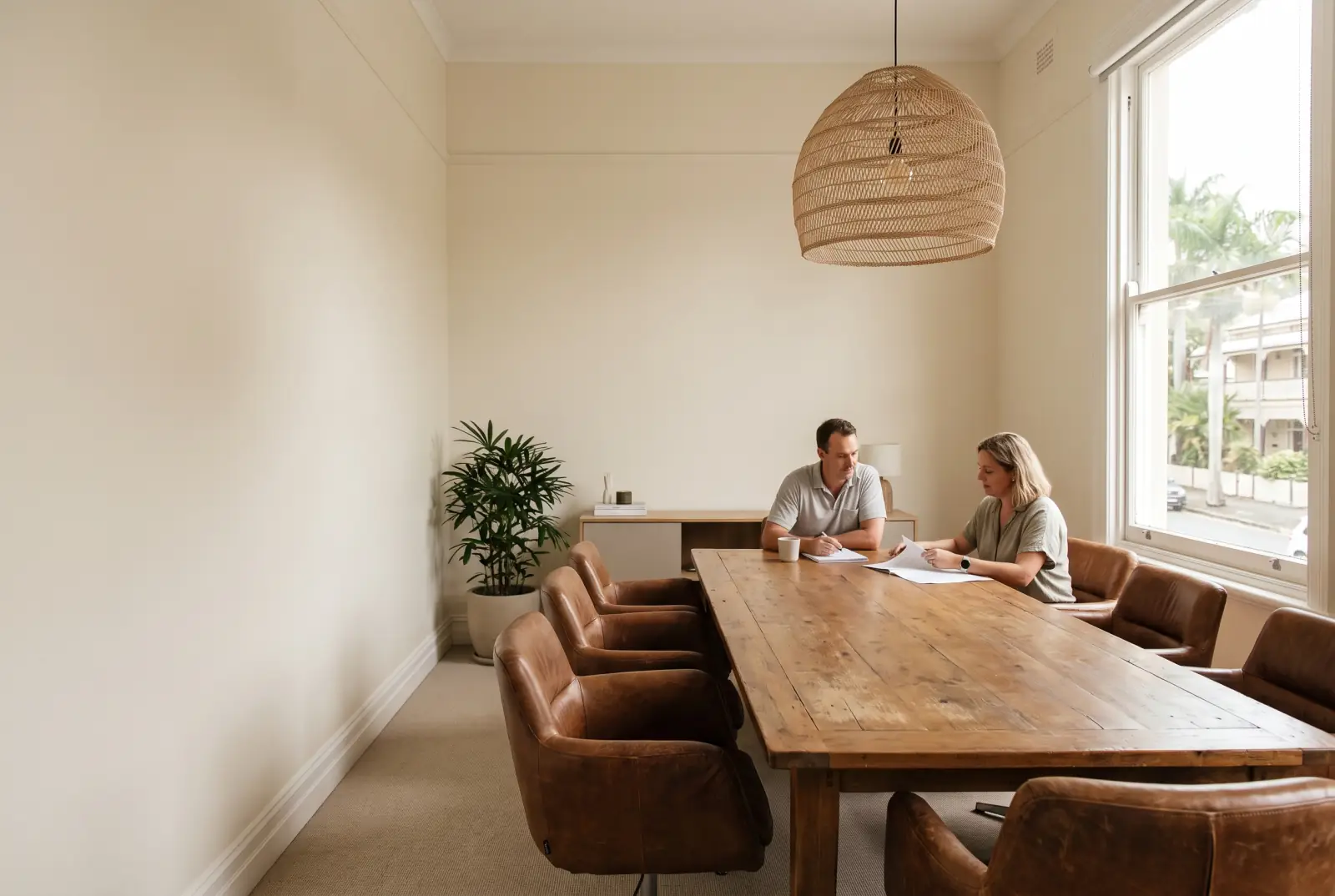 Two people meet at a long timber table in a quiet meeting room with leather chairs and a woven pendant light